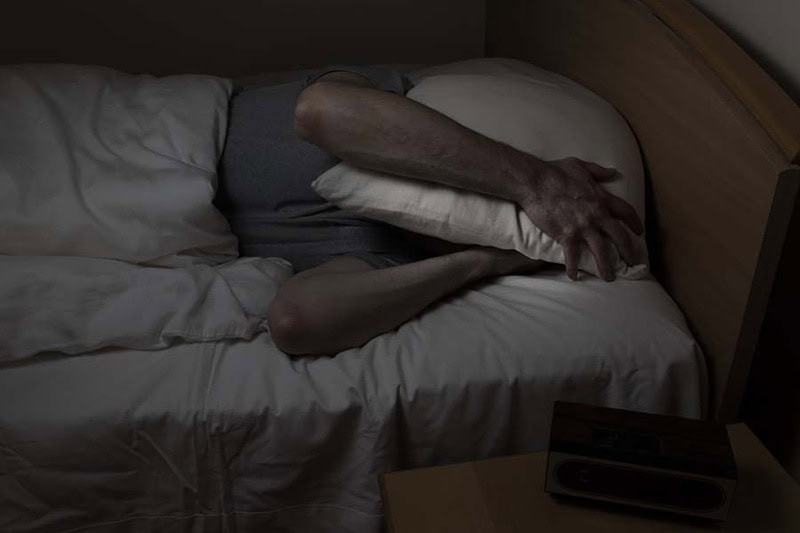 A man attempting to sleep in a bed and trying to determine why his air conditioner is making noise.