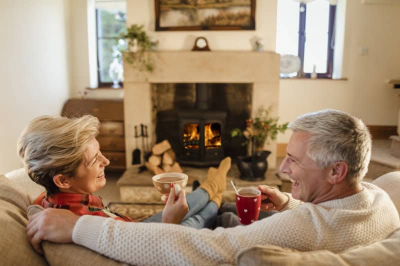 How Do I Keep My Heat During Extreme Cold Weather? - Mature couple sat infront of a fire drinking hot chocolate from mugs.