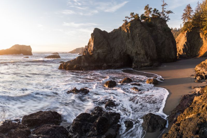 Sunset over a rocky coastal shoreline with waves crashing on dark boulders and a sandy beach area to the right