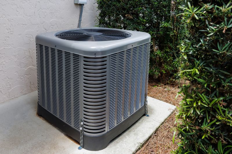 Outdoor central air conditioner condenser unit on a concrete pad beside a stucco wall with shrubs nearby