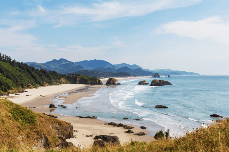 Sunlit sandy beach with rocky outcrops along a blue ocean and distant mountains under a clear sky, on a calm day.