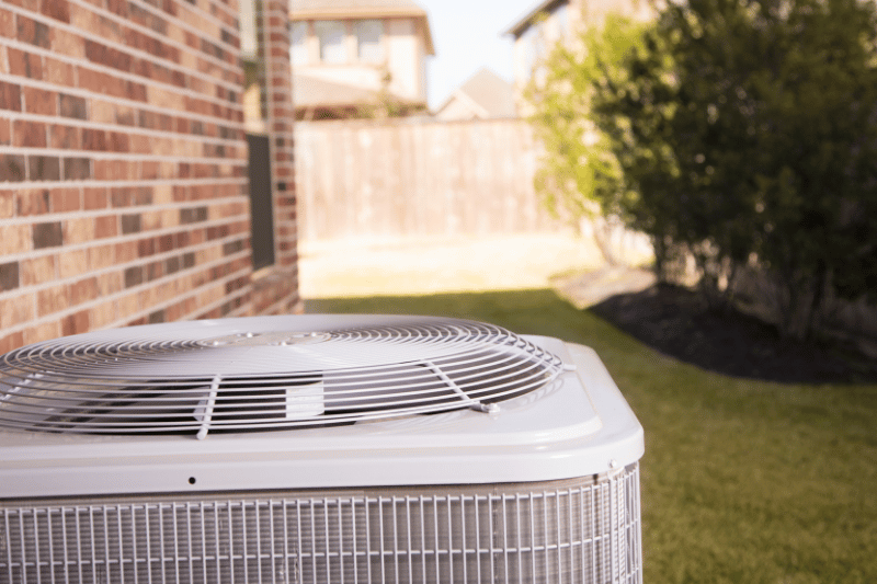 Outdoor air conditioner unit on a lawn beside a brick house with a wooden fence in the background.
