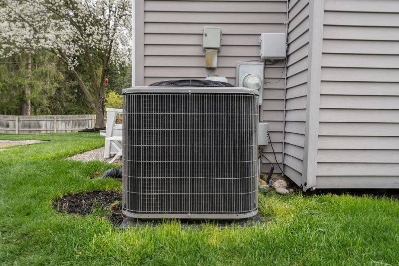 Outdoor central air conditioning condenser unit on a concrete pad beside a gray house, with a green lawn and flowering trees nearby.