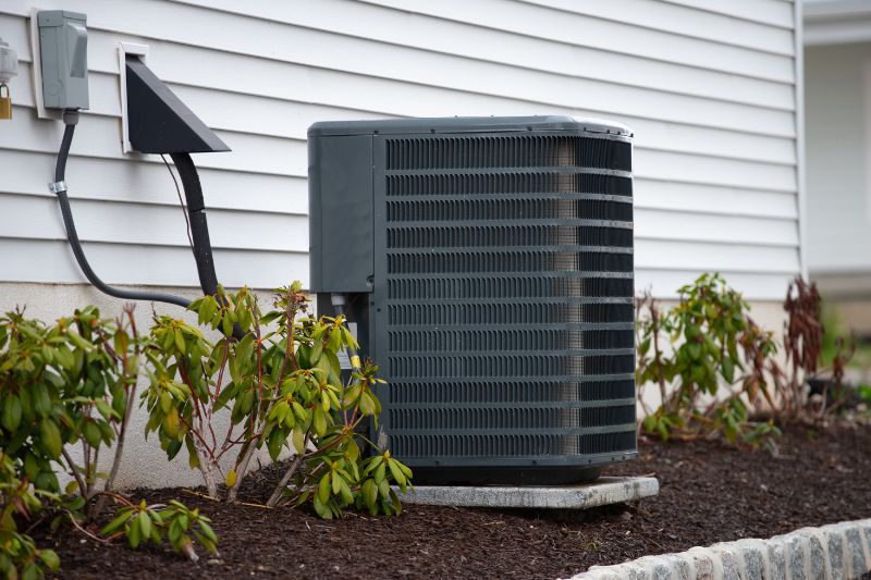 Outdoor air conditioning condenser unit on a concrete pad beside the house, with surrounding shrubs in a landscaped bed.