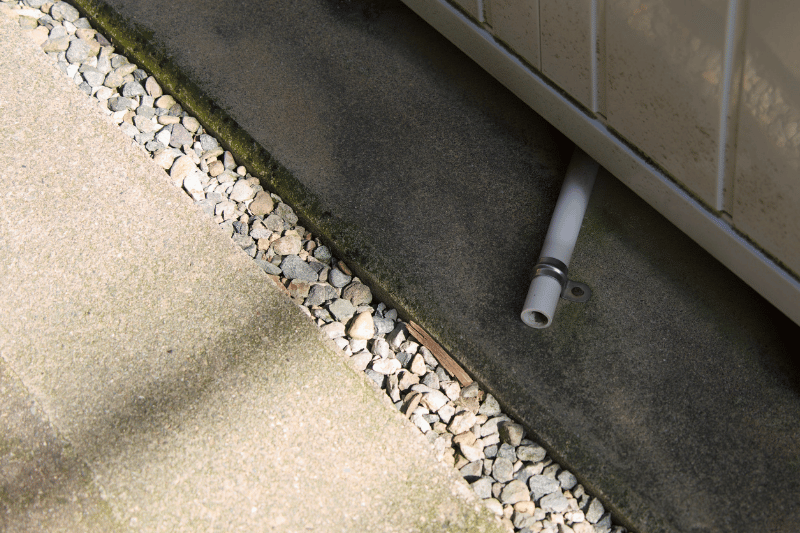 Grey downspout pipe protruding from a beige wall, resting on a concrete edge beside a gravel strip and stepping stones.