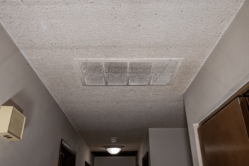 Interior hallway with a textured ceiling, rectangular ceiling vent, and doors along the corridor, lit by a ceiling light on the far end.